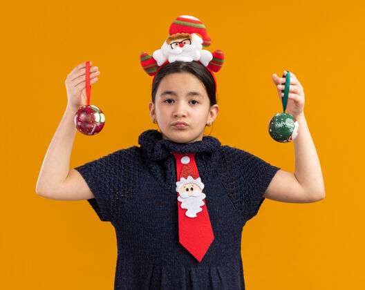 搞笑小女孩穿着针织连衣裙 头上系着一条红色领带 头上戴着一个滑稽的边 手里拿着圣诞球 严肃的脸站在橙色的背景上看着摄像机快乐女孩头