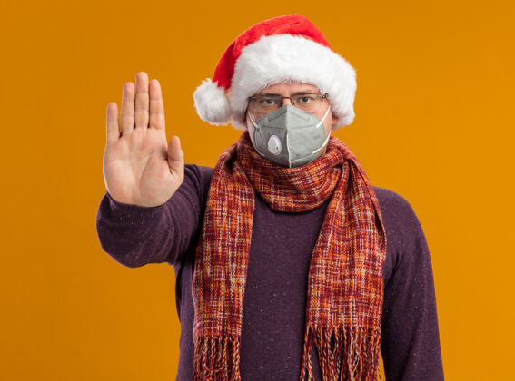 圣诞快乐严肃的成年男子戴着防护面具眼镜 戴着圣诞帽 脖子上围着围巾 看着摄像机做着停止手势 孤立在橙色背景上做手势认真