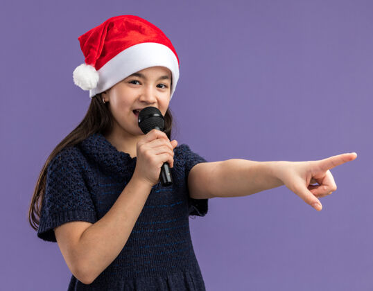 快乐小女孩穿着针织裙 戴着圣诞帽 拿着麦克风对着它说话 用食指指着紫色背景上站着的快乐的东西衣服手指编织