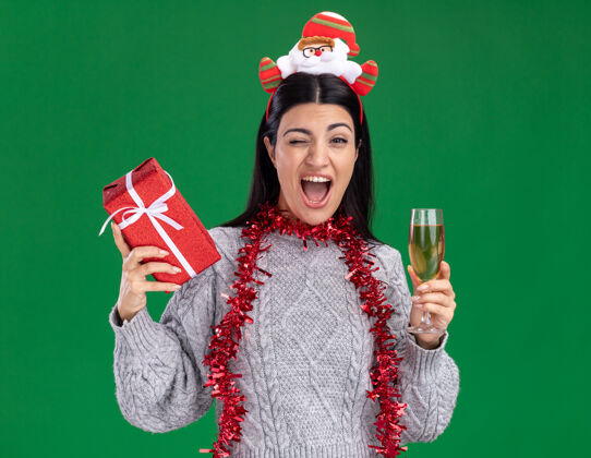 包装快乐的年轻白人女孩戴着圣诞老人的头带 脖子上戴着金箔花环 手里拿着礼包和一杯香槟 在绿色的墙上眨眼花环玻璃圣诞节