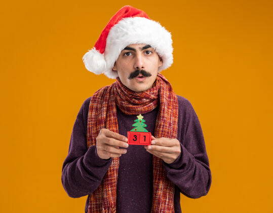 温暖一个留着胡子的男人戴着圣诞老人帽 脖子上围着暖和的围巾 手里拿着新年礼物的玩具立方体 站在橙色背景下焦急地看着相机圣诞节举行帽子