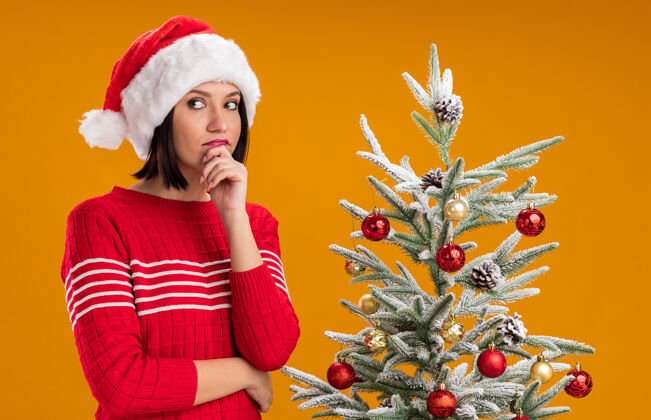 穿着体贴的年轻女孩戴着圣诞帽站在装饰好的圣诞树旁 手放在下巴上 看着橘色墙上孤立的一面年轻圣诞节靠近