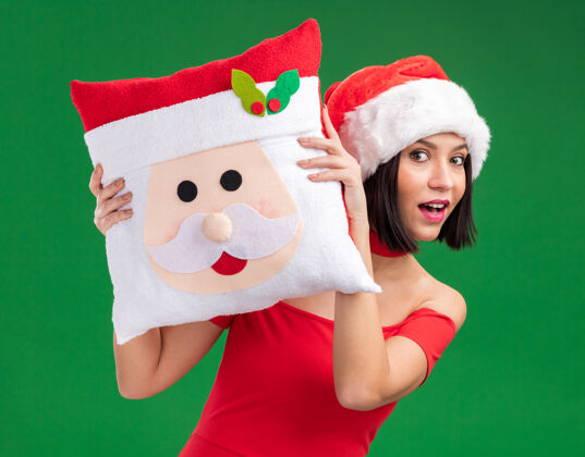 圣诞节印象深刻的年轻女孩戴着圣诞帽 抱着圣诞老人的枕头 从后面把它隔离在绿色的墙上枕头圣诞老人圣诞老人