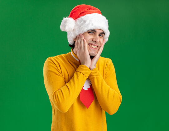 圣诞节一个穿着黄色高领毛衣 戴着圣诞帽 打着滑稽领带的年轻人站在绿色的墙上 脸上洋溢着愉快的笑容圣诞老人男人年轻人