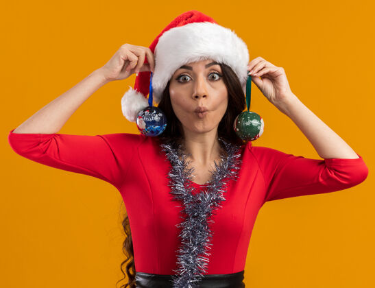 圣诞饰品顽皮的年轻漂亮女孩戴着圣诞帽 脖子上戴着金箔花环 耳朵上挂着圣诞饰品 看着相机 把鱼脸孤立在橙色背景上周围耳朵帽子