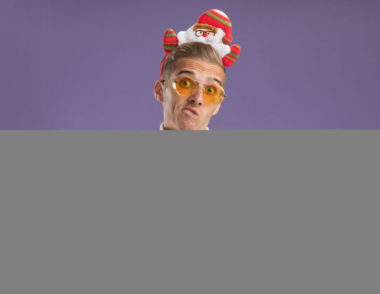 领带笨手笨脚的年轻帅哥戴着圣诞老人的头带和领带戴着眼镜拿着圣诞球饰品孤立在紫色的墙上留着复制空间圣诞老人饰品头带