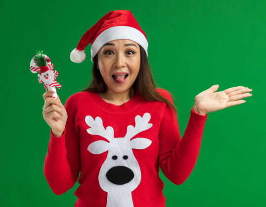 舌头年轻女子戴着圣诞老人帽 穿着红色毛衣 手持圣诞糖果手杖 看着相机 高兴而兴奋地伸出舌头站在绿色的背景上圣诞老人举行年轻