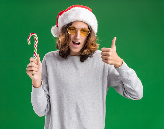 拇指快乐的年轻人戴着圣诞老人的帽子 戴着黄色的眼镜 拿着糖果手杖 看着相机 兴高采烈地微笑着 竖起大拇指站在绿色的背景上帽子年轻人眼镜
