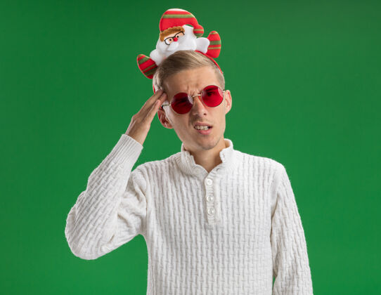头带迷茫的年轻帅哥戴着圣诞老人的头带 戴着眼镜 一边看一边摸着隔离在绿墙上的头圣诞老人年轻身边