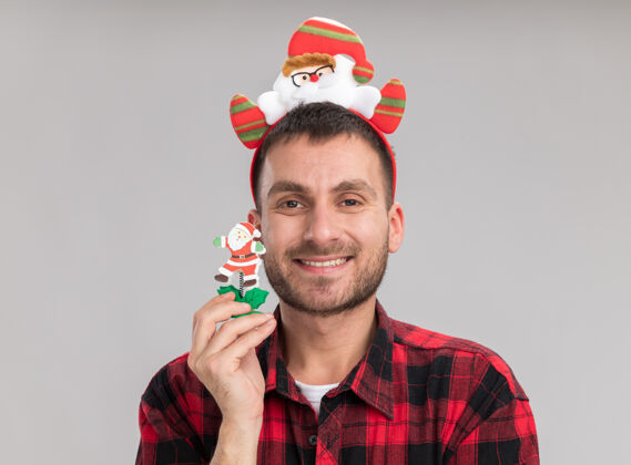 微笑微笑的白人年轻人戴着圣诞老人的头带拿着雪人的圣诞玩具看着隔离在白色背景上的相机圣诞老人抱着头带