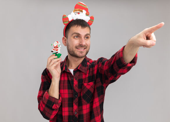 圣诞老人微笑的白人年轻人戴着圣诞老人的头带 手里拿着雪人圣诞玩具 看着并指着白色背景上孤立的一面指着雪人微笑