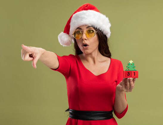 女孩令人印象深刻的年轻漂亮女孩戴着圣诞帽和眼镜拿着圣诞树玩具与日期看 并在橄榄绿的背景上指着一边孤立圣诞圣诞老人圣诞树