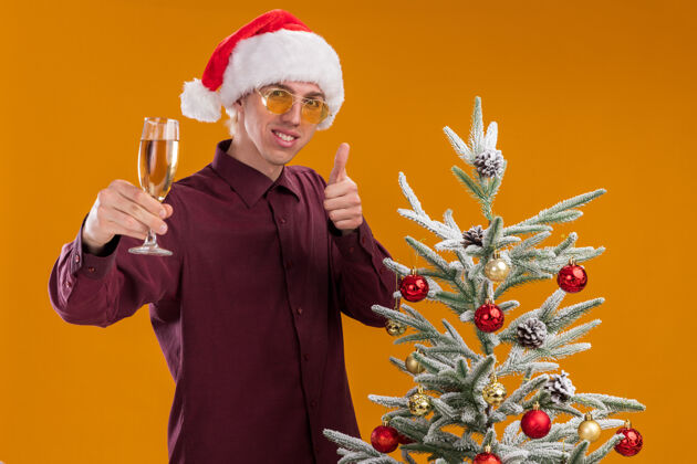 橙色带着圣诞帽 戴着圣诞眼镜 面带微笑的金发年轻人站在装饰好的圣诞树旁 伸出一杯香槟伸展圣诞快乐金发
