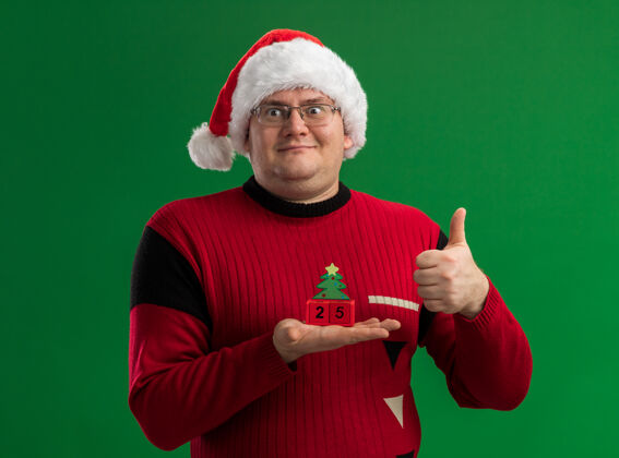 高兴戴着眼镜 戴着圣诞帽 手里拿着圣诞树玩具的快乐的成年男子 一边看一边在绿色背景上竖起大拇指圣诞老人成人帽子