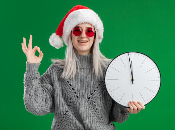 微笑年轻的金发女郎 穿着冬衣 戴着圣诞帽 戴着红眼镜 拿着挂钟 看着相机 兴高采烈地微笑着 绿色背景上立着ok标志站着时钟年轻