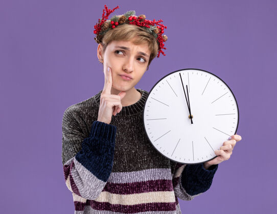 圣诞节体贴的年轻漂亮女孩戴着圣诞花环拿着时钟手放在下巴上看着紫色背景上孤立的一面侧抱着看