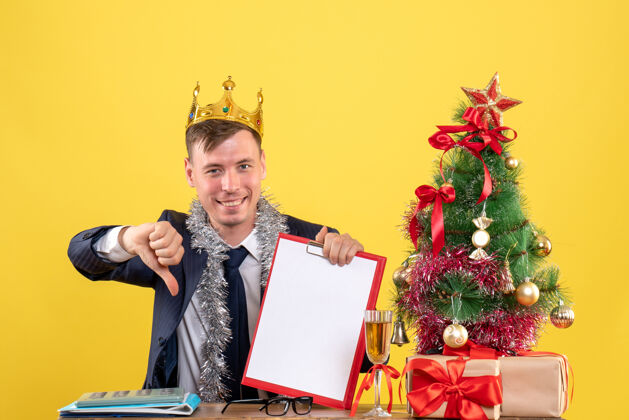 制作正面图微笑的商人做拇指向下的标志坐在圣诞树附近的桌子上 黄色的礼物电脑笔记本电脑男人