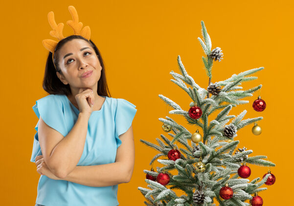鹿身着蓝色上衣的快乐年轻女子 戴着滑稽的镶边和鹿角 站在橙色背景下的圣诞树旁 抬头思考着积极的一面橘子站角
