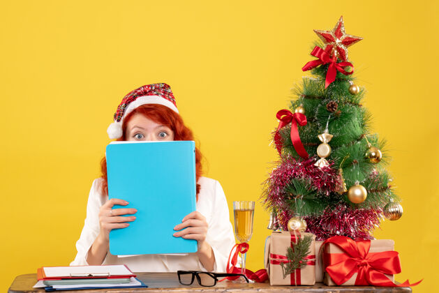 礼物正面图：女医生坐在桌子后面 拿着黄色背景的文件 还有圣诞树和礼品盒抱着坐着背景