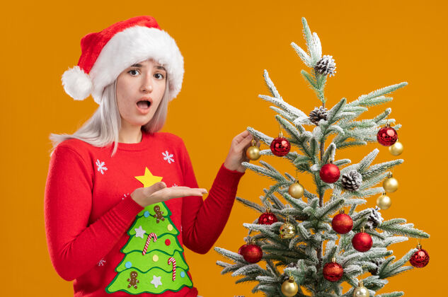 困惑穿着圣诞毛衣 戴圣诞帽的金发女郎站在一棵圣诞树旁 手挽着手在橙色背景上展示圣诞树站着帽子介绍