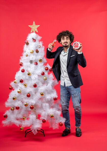 钟兴奋的年轻人拿着时钟 举起一杯葡萄酒 站在圣诞树附近的红股票照片衣服兴奋杯子