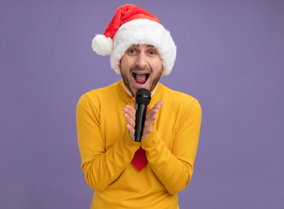抱着戴着圣诞帽打着领带的白人年轻人拿着麦克风看着摄像机在紫色背景上孤立地唱歌 还有复制空间相机帽子看