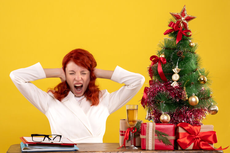 办公室女工人坐在桌子后面 手里拿着圣诞礼物 树在黄色上尖叫桌子尖叫女工人