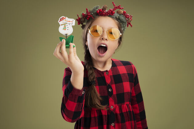 小小女孩戴着圣诞花圈 穿着格子裙 手里拿着圣诞玩具 站在绿色的背景下 高兴而兴奋地看着相机圣诞节检查兴奋