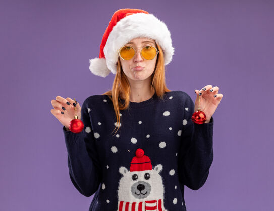 眼镜悲伤的年轻漂亮的女孩穿着圣诞毛衣和帽子戴着眼镜拿着圣诞球孤立在紫色的背景上毛衣女孩圣诞节