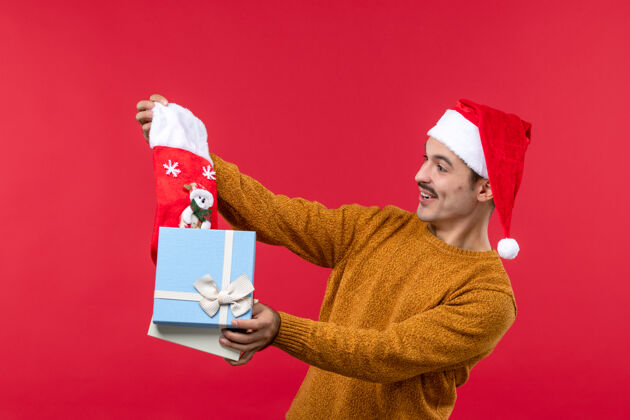 微笑年轻人在红墙上拿出圣诞袜的正面图帽子商务节日