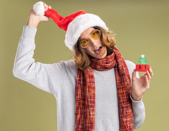 帽子年轻人戴着圣诞老人帽 戴着黄色眼镜 脖子上围着暖和的围巾 手里拿着新年快乐的玩具方块 伸出舌头站在绿色的背景上温暖日期周围