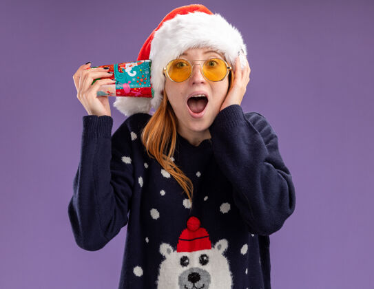 放着惊讶的年轻漂亮女孩穿着圣诞毛衣戴着圣诞帽戴着眼镜耳朵上拿着圣诞杯手放在脸颊上紫色背景上隔离毛衣脸颊年轻