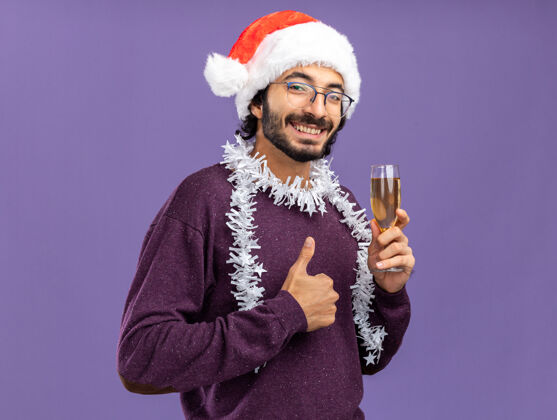 微笑微笑着的年轻帅哥戴着圣诞帽 脖子上戴着花环 手里拿着一杯香槟 在蓝色背景上孤立地竖起大拇指拇指香槟展示