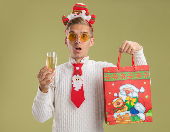 圣诞老人令人印象深刻的年轻帅哥戴着圣诞老人的头带和领带拿着一杯香槟和圣诞礼包隔离在橄榄绿的墙上领带年轻圣诞节
