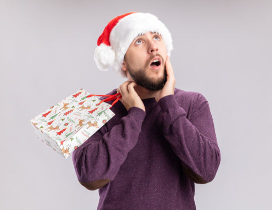 帽子穿着紫色毛衣 戴着圣诞老人帽的年轻人拿着礼品纸袋 站在白色背景上困惑地抬头看年轻圣诞老人礼物