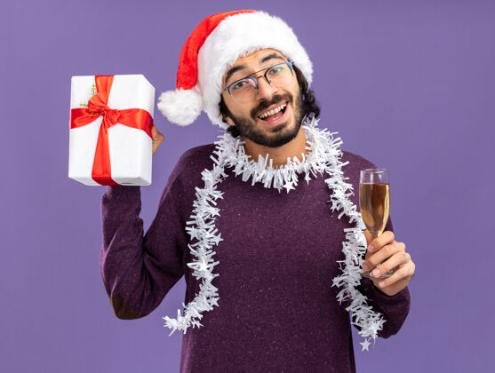 穿着兴奋的年轻帅哥戴着圣诞帽 脖子上戴着花环 手里拿着礼品盒 蓝色的墙上隔着一杯香槟帽子花环帅气