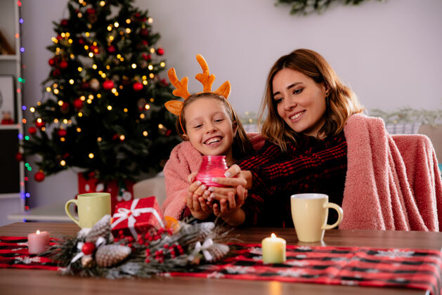 时间笑眯眯的母女俩捧着蜡烛望着桌子坐在家里享受圣诞节的时光餐桌坐着圣诞节
