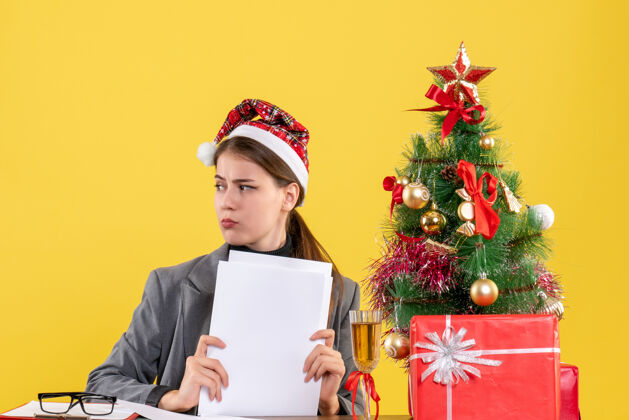 电脑前视图体贴的女孩戴着圣诞帽坐在桌子旁看着右边的圣诞树和礼物鸡尾酒鸡尾酒笔记本电脑女孩