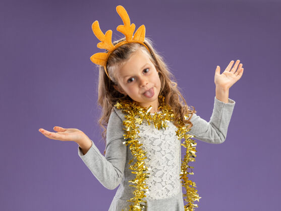 穿着快乐的小女孩戴着圣诞发箍 脖子上戴着花环 双手张开 在蓝色的墙上露出孤立的舌头小花环展示