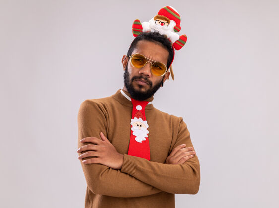 非洲非洲裔美国人 身穿棕色毛衣 头戴圣诞老人戒指 打着有趣的红色领带 眉头紧锁 双臂交叉 站在白色背景上头站立脸