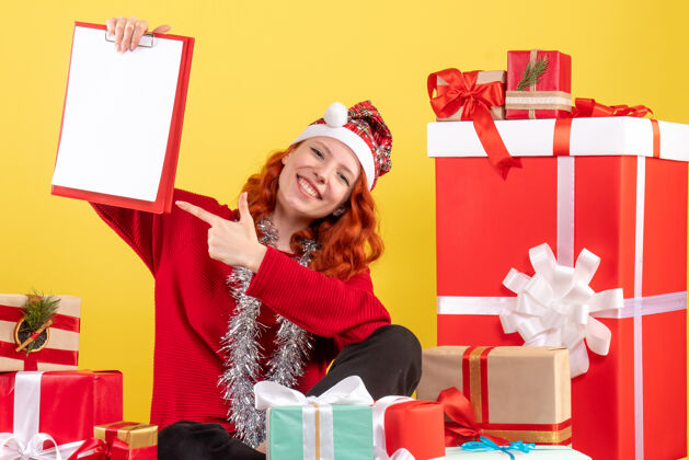 礼物坐在圣诞礼物周围的年轻女子的正面视图 黄色墙上有文件说明礼物微笑快乐