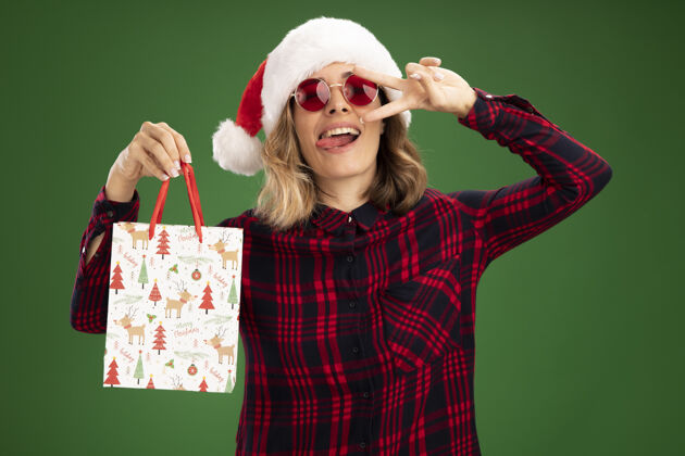 礼物快乐的年轻漂亮的女孩戴着圣诞帽戴着眼镜拿着礼品袋露出舌头在绿色背景上孤立的和平姿态女孩圣诞节举行