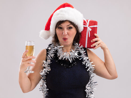 举行令人印象深刻的中年妇女戴着圣诞帽 脖子上戴着金箔花环 肩上扛着圣诞礼包 一杯香槟看着隔离在白色背景上的相机圣诞老人礼物周围