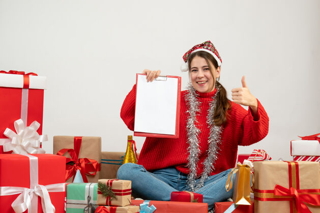 礼物戴着圣诞帽的快乐女孩拿着文件做着竖起大拇指的牌子坐在白色的礼物旁边快乐女孩盒子礼物