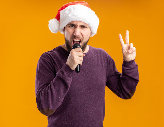 情绪化情绪化的年轻人穿着紫色毛衣 戴着圣诞帽 手持麦克风 站在橙色背景上唱歌麦克风毛衣唱歌