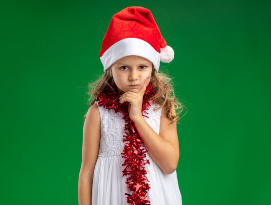 下巴皱眉的小女孩戴着圣诞帽 脖子上戴着花环 手放在下巴上 隔离在绿色的墙上放脖子小