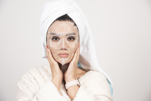 成人穿着浴衣 头戴毛巾的年轻女士的肖像 同时用面罩触摸她的脸皮肤护理浴袍面部