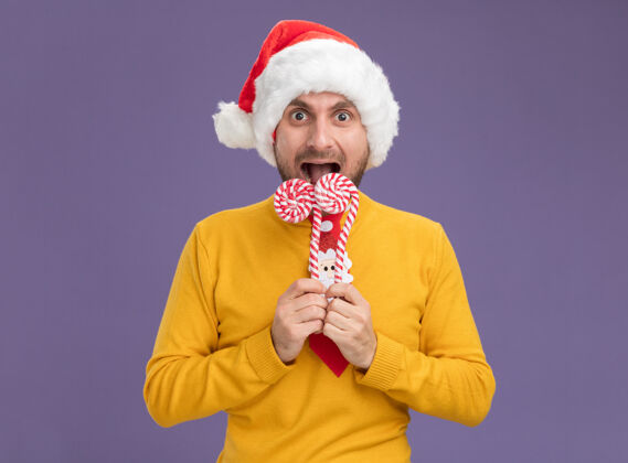 男人令人印象深刻的年轻白种人戴着圣诞帽 打着领带 拿着圣诞甜甜的手杖靠近嘴边 准备吃掉隔离在紫色墙上的手杖准备穿手杖