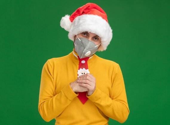 男人狡猾的白人年轻人戴着圣诞帽 打着带防护面具的领带 双手合十 在绿色的墙壁上 留着复制空间穿着狡猾年轻