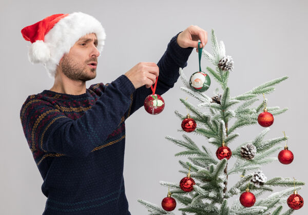 圣诞节一个戴着圣诞帽的白人年轻人站在圣诞树旁 用隔离在白色墙上的圣诞装饰球来装饰圣诞树靠近男人穿着